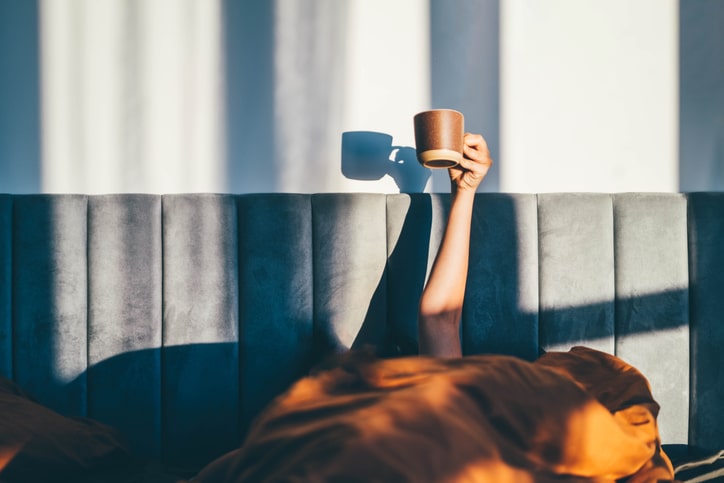 hoe overleef je een kater als moeder - vrouw steekt arm omhoog uit bed met kopje koffie